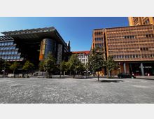 Weitläufiger Stadtplatz mit vereinzelten Bäumen im Vordergrund, flankiert von modernen Bürogebäuden aus Glas und Stahl links und einem orangefarbenen Gebäude mit waagerechten Linien rechts. Im Hintergrund ist ein klassisches Gebäude mit roten Dachziegeln sichtbar.
