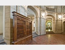 Ein historisches Innenraumfoto mit Bögen und Säulen aus hellem Stein, dekorativen Wandleuchten und einem großen Holzmöbelstück mit der Aufschrift „Allied Control Authority“. Der Boden ist mit roten Fliesen ausgelegt, und im Hintergrund sind Türen und Fensterrahmen sichtbar.