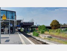 Bahnhof mit überdachtem Bahnsteig und Gleisen, im Hintergrund eine Brücke mit Graffiti und der Berliner Fernsehturm. Das Gelände ist teils bewachsen, und links sind Passagiere sowie verschiedene Bahnsteigschilder erkennbar.