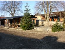 Ein ländlicher Innenhof mit Kopfsteinpflaster und mehreren Gebäuden mit orangefarbenen Fassaden und flachen Dächern. Im Vordergrund stehen Tannenbäume sowie vereinzelt Tische und Stühle unter einer überdachten Terrasse, umgeben von kahlen Laubbäumen.