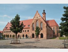 Backsteinbau eines historischen Klosters mit roten Dächern und gotischen Fensterbögen, umgeben von einem gepflasterten Hof mit vereinzelten jungen Bäumen, bei klarem Himmel. Im Hintergrund ist ein Kirchturm mit Spitzdach zu sehen.