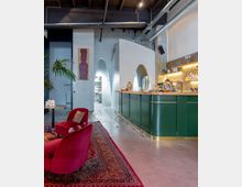 Moderne Bar in einem Loft mit sichtbaren Deckenbalken und industriellen Elementen. Im Vordergrund stehen rote Sessel auf einem orientalischen Teppich, die Bar ist grün und mit Holzplatte ausgestattet, dahinter ein offener Durchgang und Dekorationen an den Wänden.