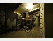 Ein altes, technisches Maschinenraum-Interieur mit sichtbaren Rohren, Metallanlagen und einem verrosteten Lüftungssystem. Die Umgebung ist spärlich beleuchtet und zeigt Betonwände sowie fest installierte Leitern.