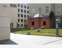 Hof eines modernen Gebäudeensembles mit einer roten, kleinen Nebengebäudestruktur mit Metalldach, umgeben von Rasenflächen, Betonpfaden und kugelförmigen Steinobjekten. Im Hintergrund Fassaden aus hellem Stein mit Fenstern.