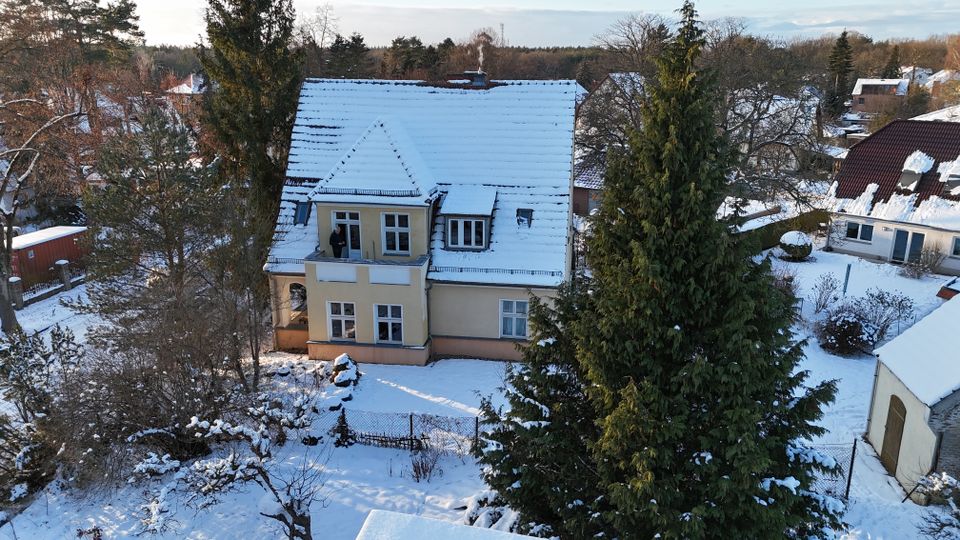 Luftaufnahme eines hellen, zweigeschossigen Wohnhauses mit steilem, schneebedecktem Satteldach und mehreren Gauben. Das Haus liegt in einem verschneiten Garten mit Zaun, umgeben von hohen Nadel- und Laubbäumen sowie benachbarten Gebäuden.