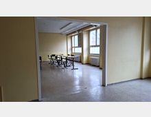 Blick durch einen breiten Durchgang in einen einfachen, leeren Klassenraum mit hellen Wänden, Linoleumboden und mehreren Schulbänken in der Mitte. Große Fenster mit Heizkörpern darunter lassen Tageslicht in den Raum; an der Decke sind lange Leuchtstofflampen montiert.