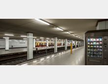Unterirdischer S-Bahnhof mit hell beleuchteten Bahnsteigen, weißen Wandfliesen, und Säulen. Auf der linken Seite fährt eine gelb-rote S-Bahn ein, rechts steht ein Getränke- und Snackautomat.