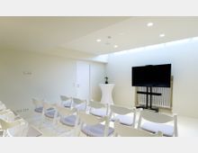 Ein kleiner, minimalistischer Konferenzraum mit weißen Wänden, mehreren weißen Stühlen in Reihen, einem schwarzen Flachbildschirm auf einem Ständer und einem Stehtisch mit einer kleinen Pflanze. Tageslicht strömt durch ein schmales Oberfenster an der Decke.