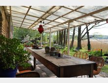 Überdachte Terrasse mit einem langen Holztisch, umgeben von Korbstühlen und dekorativen Pflanzen in Töpfen. Im Hintergrund befinden sich ein Garten mit Bäumen, blühenden Blumen und Liegestühlen sowie Schilfgras und eine Landschaft im Freien.