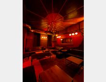 Dunkel beleuchteter Lounge- oder Barraum mit rot-oranger Lichtstimmung, schwarzen Ledersofas und mehreren niedrigen Holztischen. Sichtbare Metall-Lüftungsrohre, eine sternförmige Deckenleuchte und eine große Wandspiegel-Fläche an einer roten Akzentwand prägen den Raum.