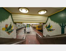 U-Bahnhof mit gelbem Zug der Linie U8 und Wandfliesen in Grün- und Weißtönen, die ein farbenfrohes Vogelmosaik darstellen. Der Bahnsteig ist mit roten und grauen Fliesen gestaltet, und ein Schild zeigt die Richtungen "Hermannstraße" und "Wittenau".