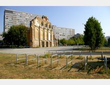 Backsteinfassade eines historischen Gebäudes, das teilweise erhalten ist, mit großen Rundbögen und Fensteraussparungen; im Hintergrund ein modernes, mehrstöckiges Wohngebäude, umgeben von Bäumen und einer asphaltierten Freifläche. Im Vordergrund befinden sich Metallbügel zur Fahrradabstellung und Grünflächen.