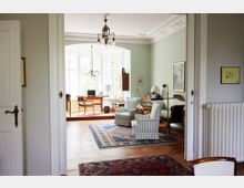 Wohnzimmer mit hohen Decken und klassischen Elementen, darunter weiße Stuckverzierungen, hellgrüne Wände und hölzerne Möbel. Im Vordergrund stehen gestreifte Sessel und Teppiche, während im Hintergrund eine Sitzecke mit Schreibtisch und großen, hellen Fenstern sichtbar ist.