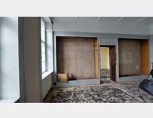 Ein leerer Raum mit abgenutztem Bodenbelag und sichtbaren Wänden, die teilweise verputzt und kahl sind. Links befinden sich große Fenster mit weißen Rahmen, und rechts ist eine unvollständige Wand mit einem offenen Durchgang zu einem weiteren Raum sichtbar.