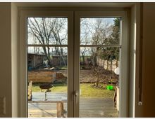 Blick durch eine Glastür auf einen Garten mit einer Holzterrasse im Vordergrund, einem Grill und einer Kerze auf einem Tisch. Im Hintergrund sind baumlose Beete, einzelne Bäume, ein Holzschuppen sowie ein Zaun zu sehen.