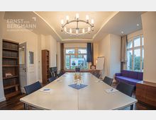 Konferenzraum mit großem rechteckigem Tisch und schwarzen Stühlen, einem zentralen Kronleuchter und einem Bildschirm am Ende des Raumes. Der Raum enthält Bücherregale, einen Kamin, ein Flipchart sowie große Fenster mit Blick ins Grüne.