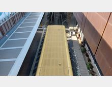 Blick auf einen Bahnsteig mit einem gelben Zug, der parallel zu den Gleisen ausgerichtet ist. Links befindet sich das überdachte Bahnsteigdach, rechts liegt eine Wand mit einer angrenzenden Schotterfläche und Müllresten.