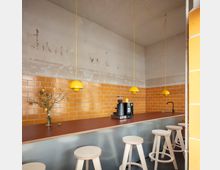 Minimalistisch gestaltete Kaffeebar mit einer Arbeitsplatte aus Holz, gelben Wandfliesen im Ziegelformat, drei hängenden gelben Lampen und hellen Holzhockern. Im Hintergrund befinden sich eine Kaffeemaschine, ein Wasserhahn und ein schlichtes Dekorationselement mit Zweigen.