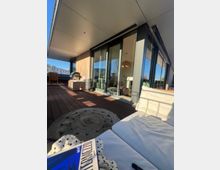 Große Dachterrasse eines modernen Wohngebäudes mit Holzdeck, breitem Dachüberstand und bodentiefen Glasschiebetüren entlang der Fassade. Auf der Terrasse stehen Sitzmöbel und ein runder Outdoor-Teppich; im Hintergrund sind umliegende Gebäude unter blauem Himmel zu sehen.