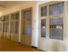 Flur in einem historischen Gebäude mit mehreren hellbeigen Holztüren, teils mit Glas- und Sprossenelementen, die in Räume führen. Der Boden ist gelblich-braun, die Wände weiß, und die Decke hat eine einfache Beleuchtung.