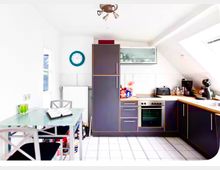 Helle Dachgeschossküche mit schrägen Decken, weißen Fliesen und L-förmiger Einbauküche in dunklen Fronten mit Holzarbeitsplatte, Herd und Spüle. Links steht ein kleiner Glastisch mit Stühlen, dazu sind Fenster und Hängeschränke an den Wänden zu sehen.