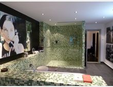 Modernes Badezimmer mit grün-schwarzen Mosaikfliesen, die die Wände und die Badewanne verkleiden. Im Raum befinden sich eine begehbare Dusche mit Glastrennwand, ein großes Kunstwerk an der Wand, sowie dunkle Fliesenböden.