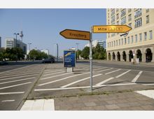 Straßenszene in Berlin mit Wegweisern zu den Stadtteilen Kreuzberg und Mitte, im Hintergrund der Fernsehturm und moderne Gebäude. Rechts ein großes, beige-farbenes Gebäude mit Rundbögen, links Bäume und eine mehrspurige Straße.