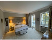 Helles Schlafzimmer mit einem Doppelbett, gepolstertem Kopfteil und zwei hängenden Lampen. Zwei große Fenster mit Blick ins Grüne, seitlich ein Bücherregal und eine weiße Heizung unter den Fenstern.