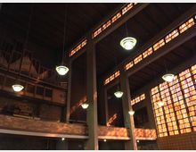 Innenraum einer modernen Kirche mit hohen Decken, sichtbaren Betonpfeilern, großen Buntglasfenstern in warmen Farben und hängenden Lampen. Im oberen Bereich befinden sich Orgelpfeifen und eine Orgelempore aus Holz.