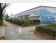 Flaches, rechteckiges Gebäude mit einer Fassade in Blau- und Grautönen, großen Fenstern und einem blauen Metalltor. Der Vordergrund zeigt einen gepflasterten Gehweg, niedrige Hecken und eine Begrenzung aus Holzstangen, umgeben von herbstlichem Laub.