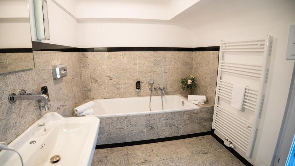 Helles Badezimmer mit grauen Steinfliesen, eingelassener Badewanne an der Rückwand und großem Spiegel über einem breiten Waschbecken. Rechts steht ein weißer Handtuchheizkörper; weiße Decke und schwarze Fliesenbordüre rahmen den Raum.