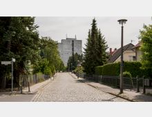 Kopfsteinpflasterstraße in einem Wohngebiet mit Einfamilienhäusern, Hecken und Bäumen an den Seiten; im Hintergrund ein mehrstöckiges Wohngebäude aus Beton. Straßenschilder und Laternen sind sichtbar.
