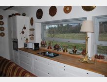 Eine helle Küche mit weißen Unterschränken und einer Holzarbeitsplatte, auf der sich Kupfergefäße, eine Kaffeemaschine, eine Mikrowelle und eine Tischlampe befinden. An der Wand hängen dekorative Kupferteller, und das Fenster gibt den Blick auf eine grüne Außenlandschaft frei.