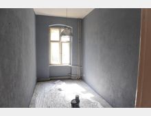 Schmaler, unbehandelter Raum mit grauen Wänden und einem einzelnen Fenster, durch das Tageslicht einfällt. Der Boden ist mit einer Schutzfolie bedeckt, und auf dem Boden steht ein Eimer mit Farbe; Rohrleitungen verlaufen entlang der Wände.