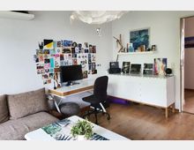 Ein heller Wohnraum mit einem Schreibtisch, auf dem ein Monitor steht, einer Wand mit einer Collage aus Fotos und Karten sowie einem weißen Sideboard mit Dekorationen und Kunstwerken. Rechts führt eine offene Tür in einen angrenzenden Raum, im Vordergrund ist ein Sofa und ein Couchtisch mit Pflanzen zu sehen.