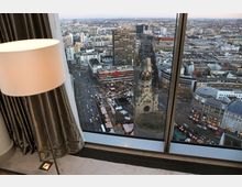 Blick aus einem hohen Gebäude auf die Kaiser-Wilhelm-Gedächtniskirche in Berlin, umgeben von einem Weihnachtsmarkt mit Ständen und Menschen. Im Hintergrund erstreckt sich die Stadtlandschaft mit modernen Gebäuden und belebten Straßen.
