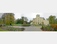 Gelbes, herrschaftliches Gebäude im klassizistischen Stil mit Turm, umgeben von einer gepflegten Gartenanlage und Bäumen; vorne eine gepflasterte Zufahrt sowie ein Parkplatz mit einem „P“-Schild.