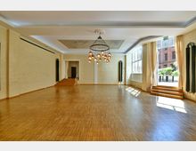 Heller Veranstaltungsraum mit Parkettboden, großen Fenstern, cremefarbenen Backsteinwänden und einem auffälligen Kronleuchter in der Deckenmitte. Der Raum verfügt über eine kleine erhöhte Bühne, blickt auf einen Außenbereich und hat einen Zugang durch Glastüren mit Vorhängen.