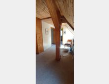 Dachzimmer mit Holzverkleidung an Wänden und Decke, sichtbarem Holzträger in der Mitte, Teppichboden sowie einer Türöffnung, die zu einem angrenzenden Raum führt. Im Hintergrund sind ein Stuhl, Bilder an der Wand und eine geöffnete Tür sichtbar.