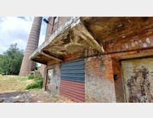 Backsteinfassade eines alten Industriegebäudes mit sichtbaren Abnutzungsspuren, teils verdeckt durch farbige Metallplatten vor einem Eingang. Im Hintergrund ist ein hoher, gemauerter Schornstein und eine Wiese mit Bäumen zu sehen.