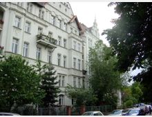 Wohnhaus im Stil des Historismus mit hellem Putz, dekorativen Fassadenelementen und Balkonen, umgeben von Bäumen und Sträuchern; am Straßenrand parken Autos.