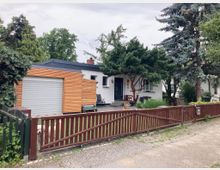 Einfamilienhaus mit flachem Dach und weißer Fassade, rechts davon ein großer Baum und ein Vorgarten mit Holzzaun. Eine angebaute Garage mit Holzverkleidung und geschlossenem Rolltor ist links zu sehen.