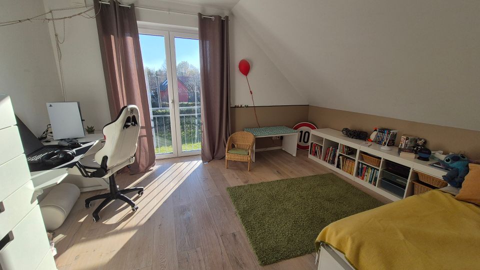 Ein modernes Kinderzimmer mit Dachschräge, großem Fenster und Terrassentür, die Tageslicht hereinlassen. Links steht ein Schreibtisch mit Computer, rechts ein Bett, Bücherregale und ein Tisch mit einem Stuhl; der Boden ist aus hellem Holz, teils mit grünem Teppich bedeckt.