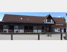 Fachwerkgebäude mit rotem Ziegeldach und Pferdeställen in der unteren Etage, davor ein gepflasterter Hof und ein Holzzaun, umgeben von blauen Himmel und grüner Vegetation im Hintergrund.
