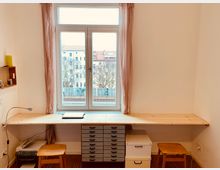 Holzarbeitsplatz vor einem großen Fenster mit rosa Vorhängen, darunter eine Schubladeneinheit, ein Drucker und zwei Holzstühle. Draußen sind Dächer und Bäume in einer urbanen Umgebung sichtbar.