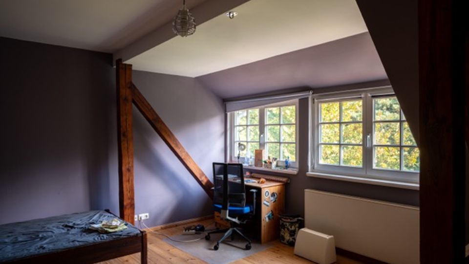Dachgeschosszimmer mit schräger Decke, Holzboden und sichtbarem Holzbalken, eingerichtet mit Bett und kleinem Schreibtisch. Drei große Sprossenfenster lassen Tageslicht herein und zeigen Bäume draußen.