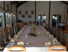 Ein festlich gedeckter langer Tisch in einem hellen Raum mit hohen Decken und Holzbalken. Der Tisch ist mit weißen Tischdecken, gefalteten Servietten, Geschirr und einem Blumengesteck dekoriert; im Hintergrund sind große Fenster und Türen sowie Bilder an den Wänden.