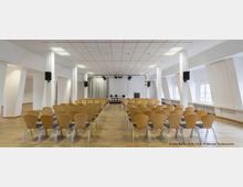 Veranstaltungsraum mit Reihen aus hellbraunen Holzstühlen, einer kleinen Bühne im Hintergrund mit vier Stühlen, einem Rednerpult und einem grauen Vorhang. Der Raum ist hell beleuchtet, hat eine abgehängte Decke mit Rasterleuchten und große Fenster auf der rechten Seite.