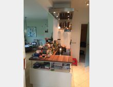 Wohnküche mit einem Holzküchenblock, der eine Arbeitsfläche mit integriertem Herd und Dunstabzugshaube bietet. Der Raum ist teils aufgeräumt, mit Küchengeräten, Hängelampen und offenen Regalen; im Hintergrund sind ein Wohnzimmer mit Sofa und Esstisch erkennbar.