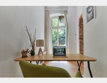 Arbeitsbereich in einem hellen Raum mit einem Holztisch, darauf ein Laptop, eine Tischlampe und Pflanzen. Im Hintergrund befindet sich ein großes Fenster mit Blick auf grüne Bäume, das von einem Backsteinbogen und weißen Wänden umrahmt wird.